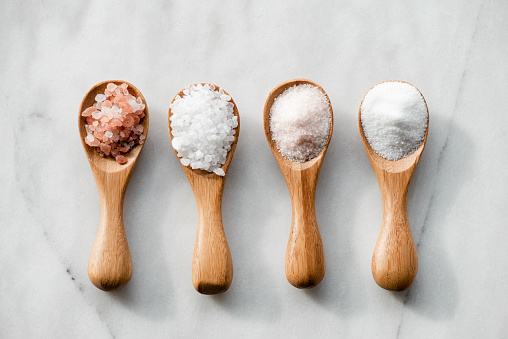 Various salt types in 4 wooden spoons on white marble.